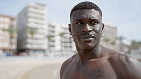 Téléchargez les photos : Africain américain touriste debout torse nu avec un visage sérieux à la plage - en image libre de droit