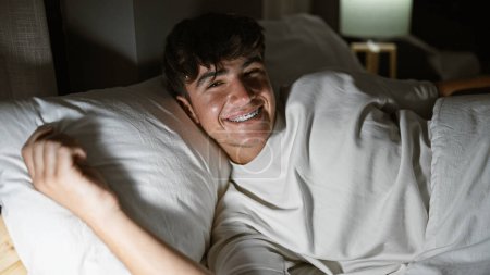 Handsome young hispanic teenager, with buoyant confidence, expressively enjoying his morning, reclining on a comfortable bed, amid the soft light in his cozy bedroom.