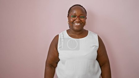 Téléchargez les photos : Femme afro-américaine confiante et souriante portant des lunettes, ayant une ambiance amusante et positive, se tenant décontractée sur un fond rose isolé. - en image libre de droit