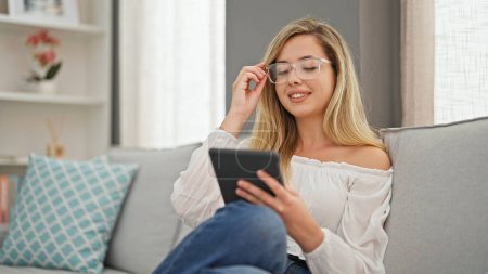 Téléchargez les photos : Jeune femme blonde portant des lunettes à l'aide de touchpad souriant à la maison - en image libre de droit