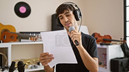 Photo for Passionate young hispanic man, a handsome musician, in the heart of a melody, reading music sheet and singing the song with a professional voice at an indoor music studio. - Royalty Free Image