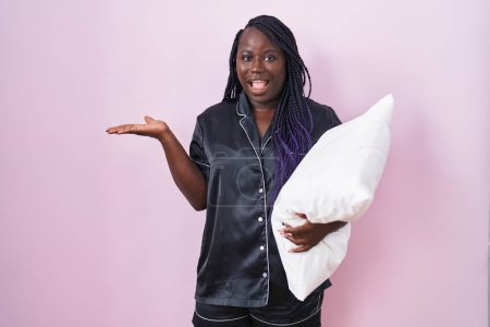 Téléchargez les photos : Jeune femme africaine portant un pyjama étreignant l'oreiller pointant de côté avec les mains ouvertes paumes montrant l'espace de copie, présentant la publicité souriant excité heureux - en image libre de droit