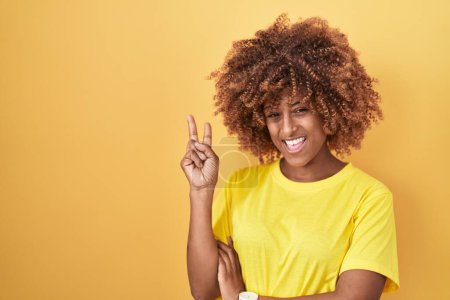 Téléchargez les photos : Jeune femme hispanique avec les cheveux bouclés debout sur fond jaune souriant avec le visage heureux clin d'oeil à la caméra faisant signe de victoire. numéro deux. - en image libre de droit