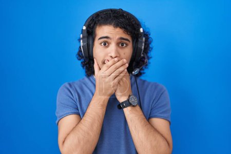 Foto de Hombre hispano con el pelo rizado escuchando música usando auriculares impactados cubriendo la boca con las manos por error. concepto secreto. - Imagen libre de derechos