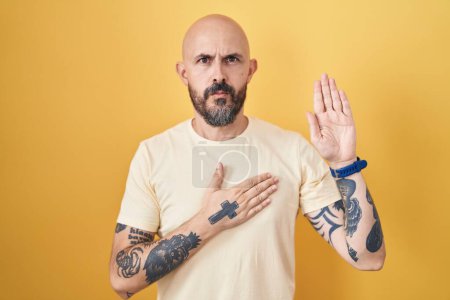Téléchargez les photos : Homme hispanique avec des tatouages debout sur fond jaune jurant avec la main sur la poitrine et la paume ouverte, faire un serment de fidélité promesse - en image libre de droit
