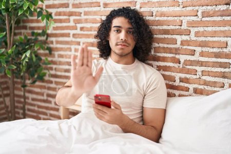 Foto de Hombre hispano con el pelo rizado usando smartphone sentado en la cama con la mano abierta haciendo stop sign con expresión seria y segura, gesto de defensa - Imagen libre de derechos