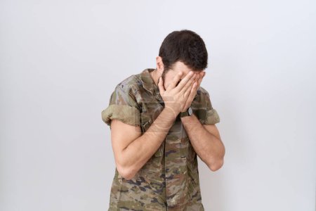Téléchargez les photos : Jeune homme hispanique portant un uniforme de camouflage de l'armée avec une expression triste couvrant le visage avec les mains tout en pleurant. concept de dépression. - en image libre de droit