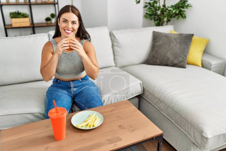 Foto de Joven hermosa mujer hispana comiendo hamburguesa sentada en el sofá en casa - Imagen libre de derechos