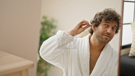 Téléchargez les photos : Jeune homme hispanique portant peignoir de nettoyage oreille avec coton-tige regardant sur miroir à la maison - en image libre de droit