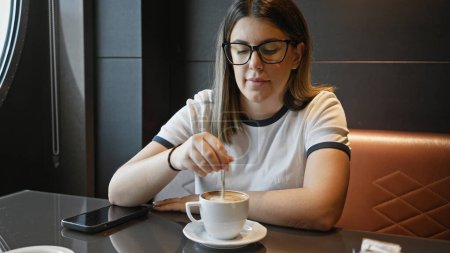 Téléchargez les photos : Une jeune femme hispanique adulte prend un café à l'intérieur d'une table de café, en remuant sa boisson de façon ciblée. - en image libre de droit