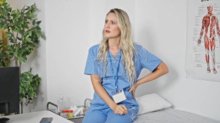 Téléchargez les photos : Jeune femme blonde médecin assis sur une table de massage avec une expression sérieuse à la clinique - en image libre de droit