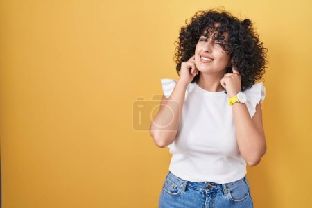 Foto de Joven mujer de Oriente Medio de pie sobre el fondo amarillo cubriendo las orejas con los dedos con expresión molesta por el ruido de la música fuerte. concepto de sordo. - Imagen libre de derechos