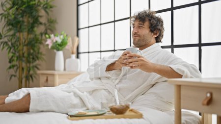 Photo for Young hispanic man wearing bathrobe holding cup of coffee at bedroom - Royalty Free Image