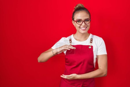 Photo for Young hispanic woman wearing waitress apron over red background gesturing with hands showing big and large size sign, measure symbol. smiling looking at the camera. measuring concept. - Royalty Free Image