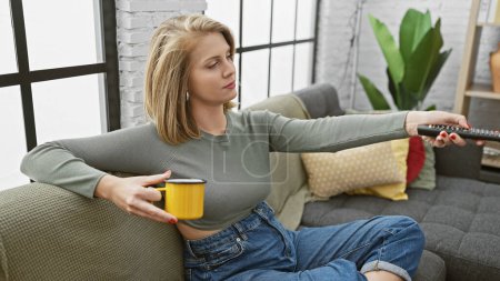 Foto de Relajada mujer rubia sosteniendo taza de café mientras usa control remoto en una acogedora sala de estar. - Imagen libre de derechos