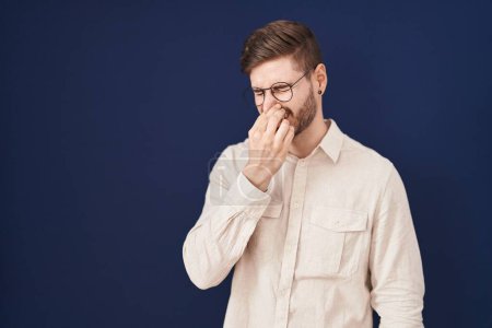 Téléchargez les photos : Homme hispanique avec la barbe debout sur fond bleu sentant quelque chose de puant et dégoûtant, odeur intolérable, retenant souffle avec les doigts sur le nez. mauvaise odeur - en image libre de droit