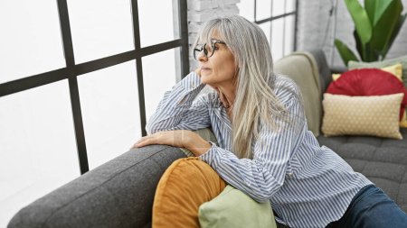 Mujer madura pensativa con el pelo gris relajándose en un sofá en un acogedor interior de la casa.