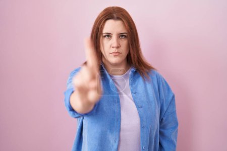 Photo for Young hispanic woman with red hair standing over pink background pointing with finger up and angry expression, showing no gesture - Royalty Free Image