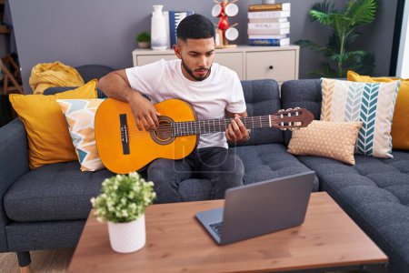 Téléchargez les photos : Jeune homme latino ayant cours de guitare classique en ligne assis sur le canapé à la maison - en image libre de droit