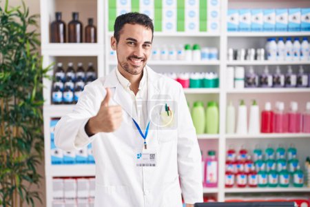 Téléchargez les photos : Un bel homme hispanique qui travaille à la pharmacie en train de faire un geste heureux du pouce à la main. approuvant l'expression regardant la caméra montrant le succès. - en image libre de droit