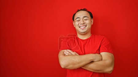 Foto de Riéndose guapo joven asiático hombre exuda confianza, de pie con brazos cruzados sobre rojo fondo, encarnando alegría y positividad - Imagen libre de derechos