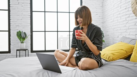 Foto de Joven mujer hispana hermosa haciendo videollamada con portátil sentado en la cama bebiendo café en el dormitorio - Imagen libre de derechos