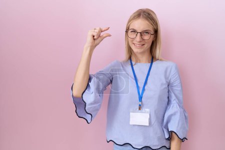 Téléchargez les photos : Jeune femme d'affaires caucasienne portant carte d'identité geste souriant et confiant avec la main faisant signe de petite taille avec les doigts à la recherche et la caméra. concept de mesure. - en image libre de droit