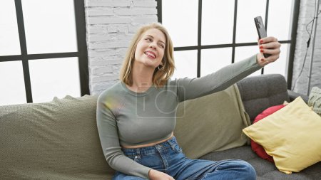 Téléchargez les photos : Une jeune femme joyeuse prend un selfie avec un smartphone sur un canapé dans un salon lumineux. - en image libre de droit