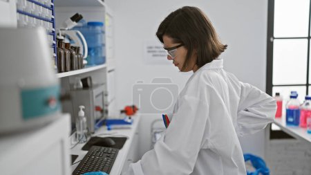 Foto de Científica hispana con exceso de trabajo al final de su ingenio, la hermosa mujer sufre de dolor de espalda por horas encorvada sobre la computadora en el laboratorio - Imagen libre de derechos