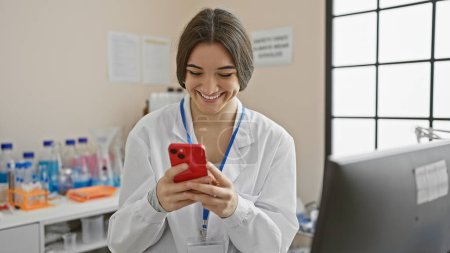Téléchargez les photos : Femme hispanique en laboratoire sourit en utilisant un smartphone, capturant un moment de professionnalisme de la santé moderne. - en image libre de droit