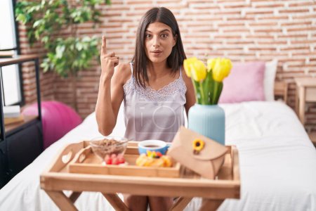 Téléchargez les photos : Brunette jeune femme manger le petit déjeuner assis sur le lit pointant du doigt avec une idée réussie. Sorti et heureux. numéro un. - en image libre de droit