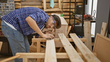 Foto de Guapo, serio, joven árabe trabajando diligentemente, lijando tablón de madera en el corazón de su ocupado taller de carpintería interior - Imagen libre de derechos