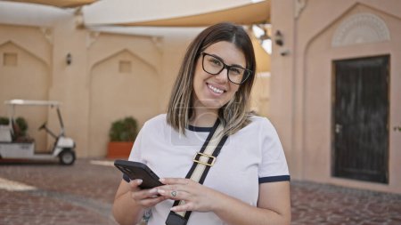 Téléchargez les photos : Jeune femme souriante avec des lunettes utilisant un smartphone dans la rue doha - en image libre de droit