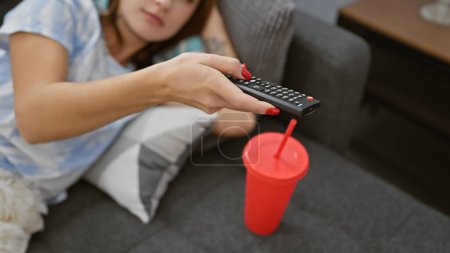 Foto de Una mujer joven descansa casualmente en un sofá en un apartamento con un mando a distancia con una taza roja en la mesa auxiliar. - Imagen libre de derechos