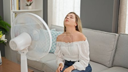Foto de Joven mujer rubia sentada en el sofá sufriendo por el calor usando ventilador en casa - Imagen libre de derechos