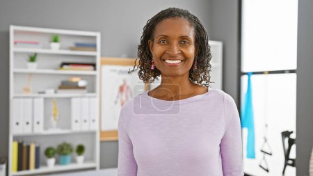 Foto de Sonriente mujer afroamericana de mediana edad en el interior de una clínica con equipo médico. - Imagen libre de derechos