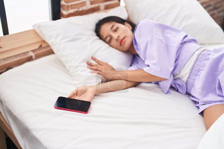 Junge schöne hispanische Frau hält Smartphone auf dem Bett liegend und schläft im Schlafzimmer
