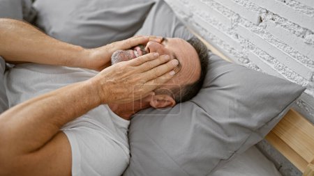 Téléchargez les photos : Homme d'âge moyen aux cheveux gris ayant une matinée confortable, couché confortablement avec les yeux à moitié éveillés dans une chambre relaxante, frottant ses yeux sur un lit couvert d'oreiller dans son appartement intérieur - en image libre de droit