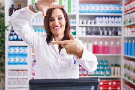 Téléchargez les photos : Femme brune d'âge moyen travaillant à la pharmacie pharmacie souriant faisant cadre avec les mains et les doigts avec le visage heureux. concept de créativité et photographie. - en image libre de droit
