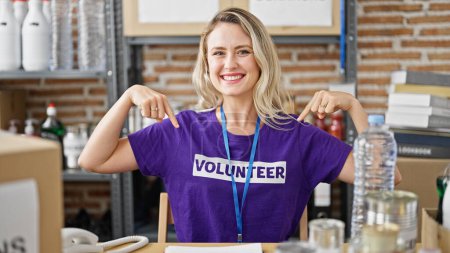 Photo for Young blonde woman sitting on table pointing herself to volunteer uniform at charity center - Royalty Free Image