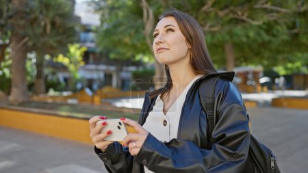 Photo for Young beautiful brunette woman holding a smartphone looks upward on a sunny urban street lined with trees. - Royalty Free Image