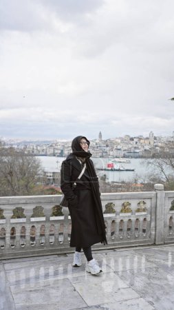 Foto de Una mujer sonriente con un abrigo negro posa con el horizonte de Estambul en el fondo, capturando viajes y exploración cultural. - Imagen libre de derechos