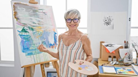 Photo for Mature woman artist smiling in a studio with a colorful painting, palette, and glasses. - Royalty Free Image