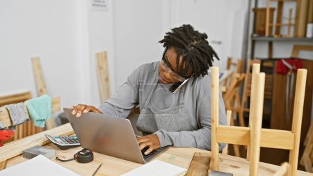 Téléchargez les photos : Jeune femme noire avec dreadlocks multitâche dans un atelier de menuiserie, en utilisant un ordinateur portable tout en parlant au téléphone. - en image libre de droit