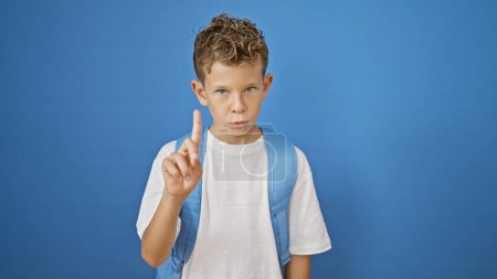 Photo for Adorable blond boy student, a smart little kid standing, serious face expressing negation, saying no with his finger. an isolated blue wall cutout background accentuates his academic bag. - Royalty Free Image