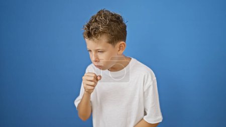 Photo for Adorable blond boy, a little sick kid, seriously coughing with flu symptoms over a blue isolated background. child's illness captured as he stands casually, concentrated on his illness. - Royalty Free Image