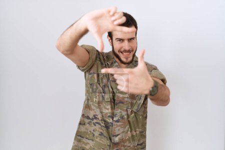 Foto de Joven hombre hispano vistiendo uniforme camuflaje del ejército sonriendo haciendo marco con manos y dedos con cara feliz. concepto de creatividad y fotografía. - Imagen libre de derechos