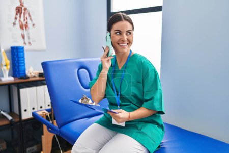 Photo for Young beautiful hispanic woman physiotherapist reading document talking on smartphone at rehab clinic - Royalty Free Image