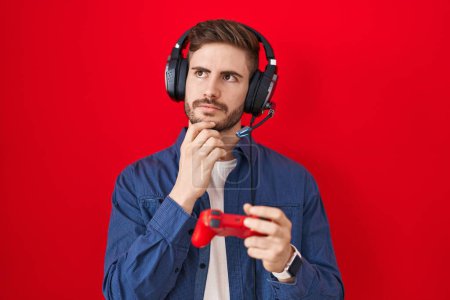 Photo for Hispanic man with beard playing video game holding controller serious face thinking about question with hand on chin, thoughtful about confusing idea - Royalty Free Image