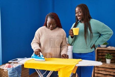 Photo for African american women ironing clothes drinking coffee at laundry room - Royalty Free Image